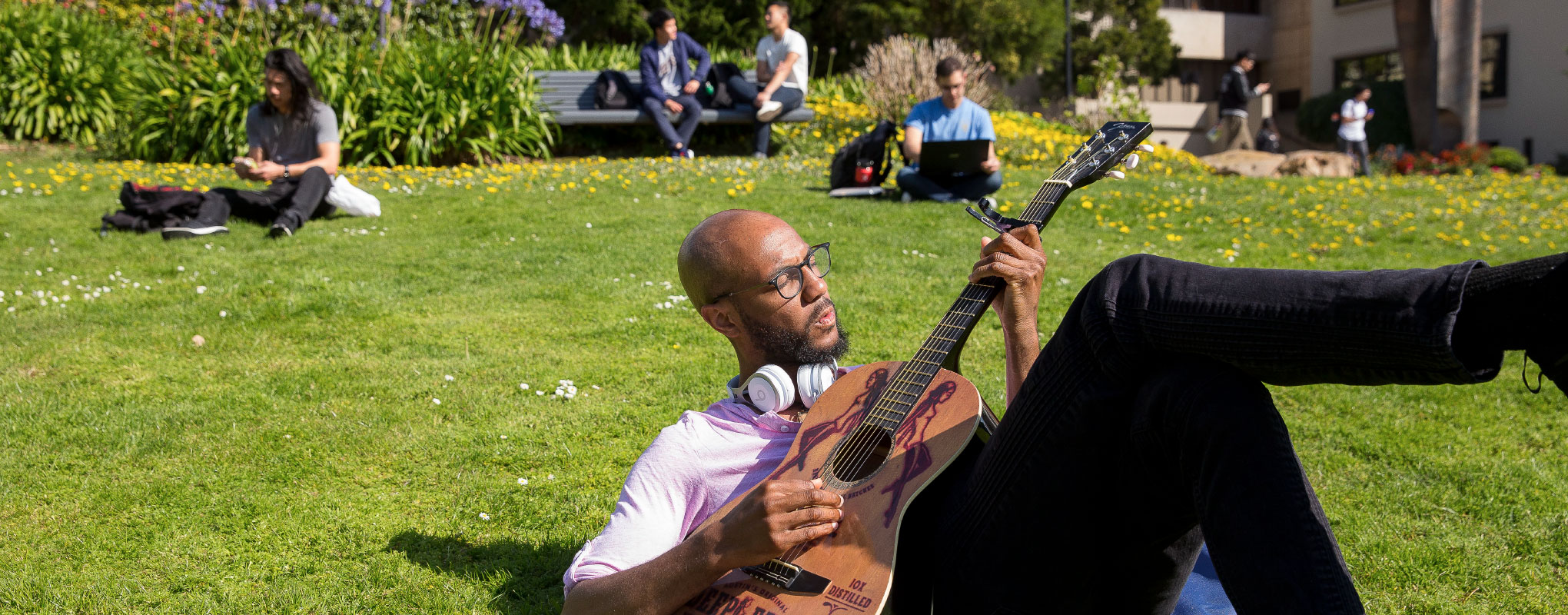 student playing their guitar out on the lawn with other students on their laptops